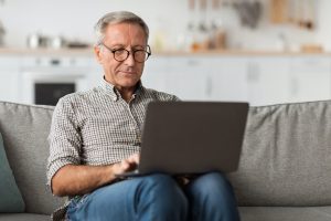 Senior Male Using Laptop Sitting On Couch At Home signing up during Medicare General Enrollment Period